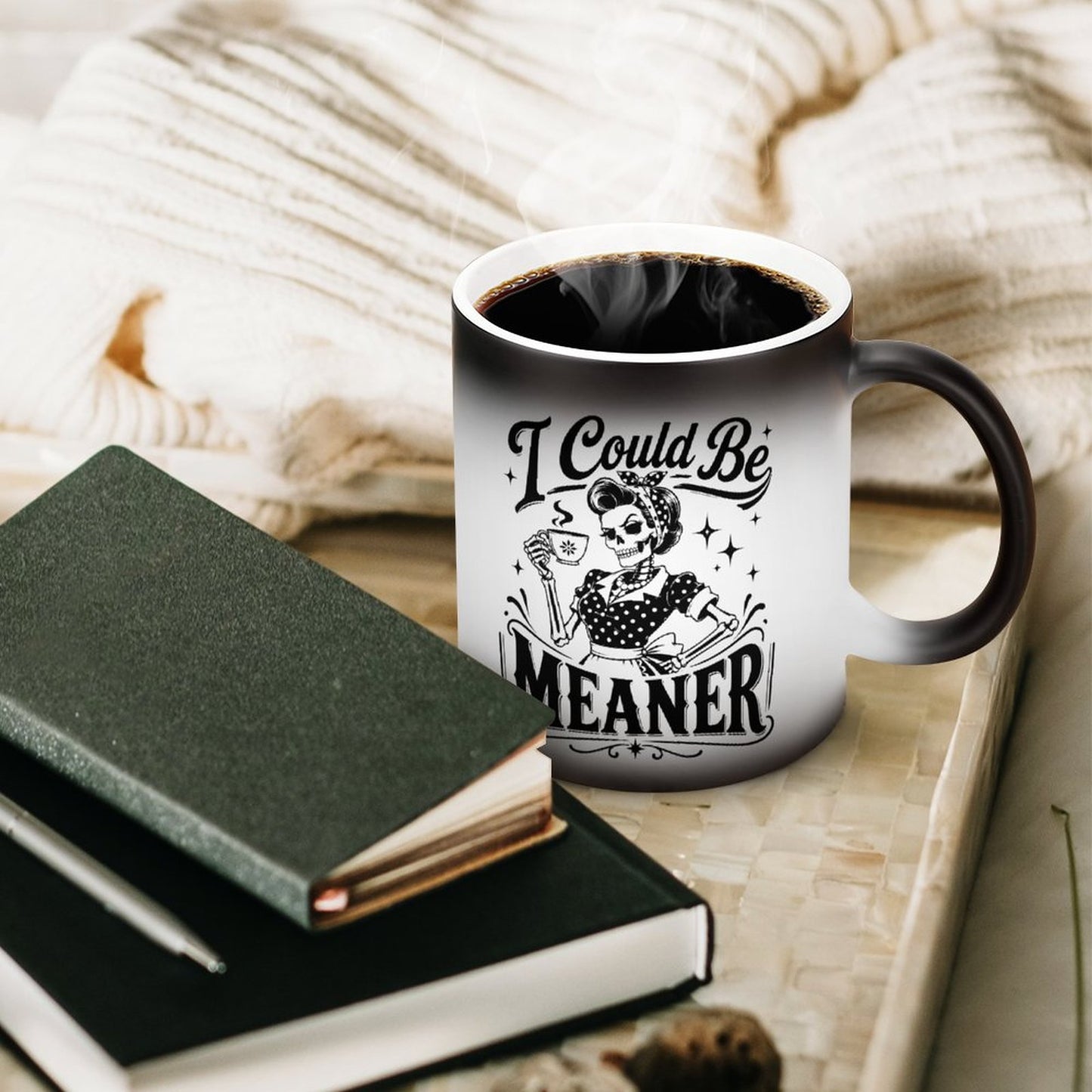 A white ceramic magic mug displaying a funny dark romance gothic skeleton design with the quote I Could Be Meaner next to a notebook.