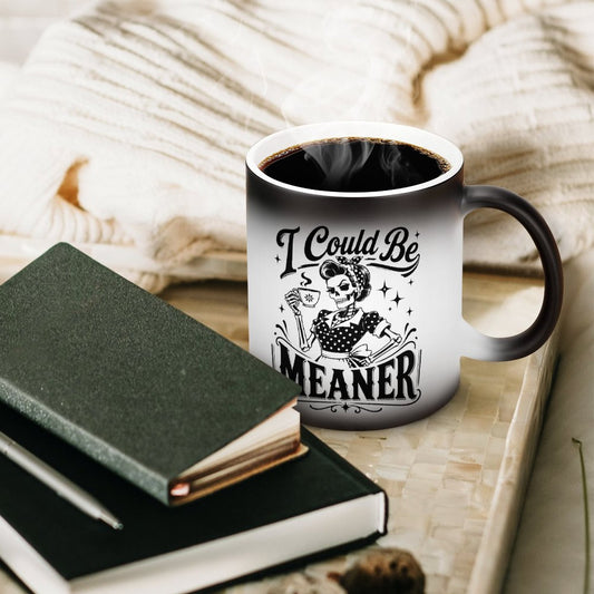A white ceramic magic mug displaying a funny dark romance gothic skeleton design with the quote I Could Be Meaner next to a notebook.