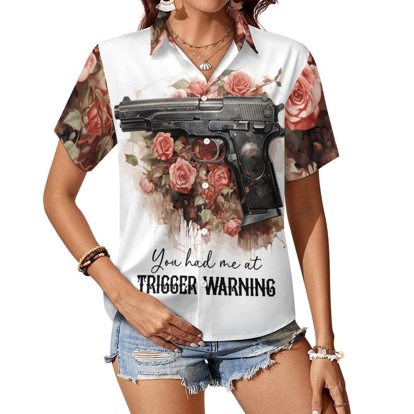 Woman wearing the 'You Had Me at Trigger Warning' short-sleeve button-down shirt paired with denim shorts. The white shirt features an all-over dark romance pattern of pink roses and a handgun graphic design on the front.